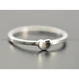 May include: A silver ring with a small, round bead at the center. The ring band is thin and polished, and the bead is smooth and reflective. The ring is set against a gray background.