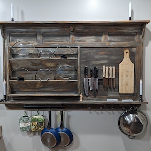 May include: A rustic wooden kitchen organizer with a shelf for storing pot lids, a magnetic strip for knives, and hooks for hanging utensils. The organizer has a distressed, weathered finish and is mounted on a wall.