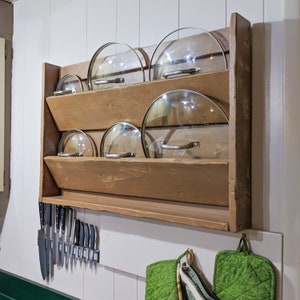 Puede incluir: Un estante de pared de madera con cinco tapas de vidrio para ollas y sartenes. El estante está teñido de un color marrón oscuro y tiene un estante debajo. También hay varios cuchillos colgados en una tira magnética en la pared.