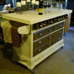 May include: A white kitchen island with a wood countertop and brown drawers. The island has a wine rack and a towel rack. The island is on wheels and has a white and brown paint finish.
