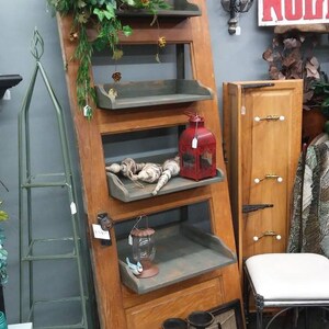May include: A repurposed wooden door transformed into a rustic shelf unit. The door features four shelves, decorated with a red lantern, decorative items, and greenery. The unit leans against a gray wall, showcasing a vintage aesthetic.