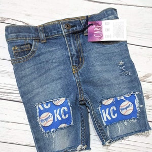 May include: Blue denim shorts with distressed details and patches featuring a baseball and the text "KC Royals".