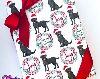 May include: White wrapping paper with a repeating pattern of black Labrador Retrievers wearing Santa hats. The dogs are surrounded by wreaths with red berries and green leaves. The text "Merry Christmas" and "Joy" are printed on the paper.