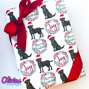 May include: White wrapping paper with a repeating pattern of black Labrador Retrievers wearing Santa hats. The dogs are surrounded by wreaths with the words "Merry Christmas" and "Joy".