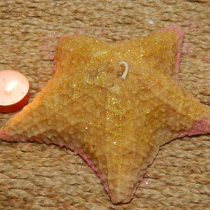May include: A starfish-shaped candle with a textured, glittery surface and pink edges. A small, lit tea light candle sits nearby. The candle is a golden yellow color and rests on a woven, brown surface.