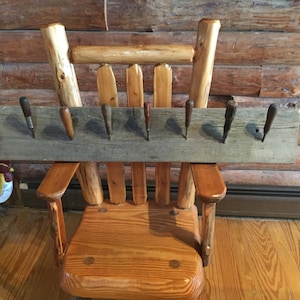 May include: A rustic wooden chair with a weathered plank attached to the back. The plank has eight metal hooks for hanging tools or other items. The chair is made of natural wood and has a simple, rustic design.