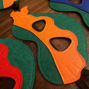 May include: Four felt masks in different colors, each with a unique design. The masks are green, orange, red, and blue. Each mask has a black eye hole and a black elastic band.