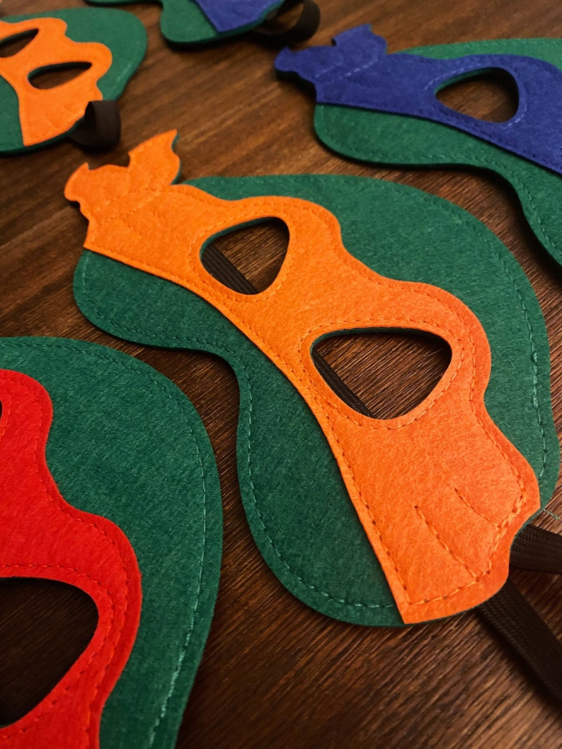 May include: Four felt masks in different colors, each with a unique design. The masks are green, orange, red, and blue. Each mask has a black eye hole and a black elastic band.