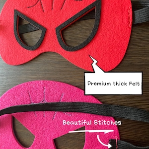 May include: Two felt superhero masks with black elastic straps. The red mask has a spider web design and the pink mask has a simple design. Both masks have black eye holes and are made with premium thick felt. The pink mask has the text "Beautiful Stitches" written on it.