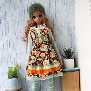 May include: A doll wearing a green hat, a blue and brown floral dress with orange trim, striped tights, and brown boots. The doll is standing in front of a light blue wooden background.