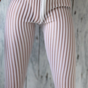 Könnte beinhalten: Nahaufnahme einer gestreiften Leggings in Weiß und Hellbraun. Die vertikalen Streifen erzeugen ein visuell interessantes Muster. Die Leggings sind Teil eines Kleidungssets, mit weißer Spitzentrimmung am Bund.