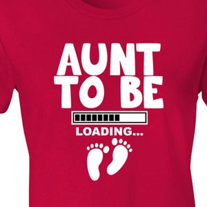 May include: Red t-shirt with white text that reads "Aunt to Be" with a loading bar and two white baby footprints.