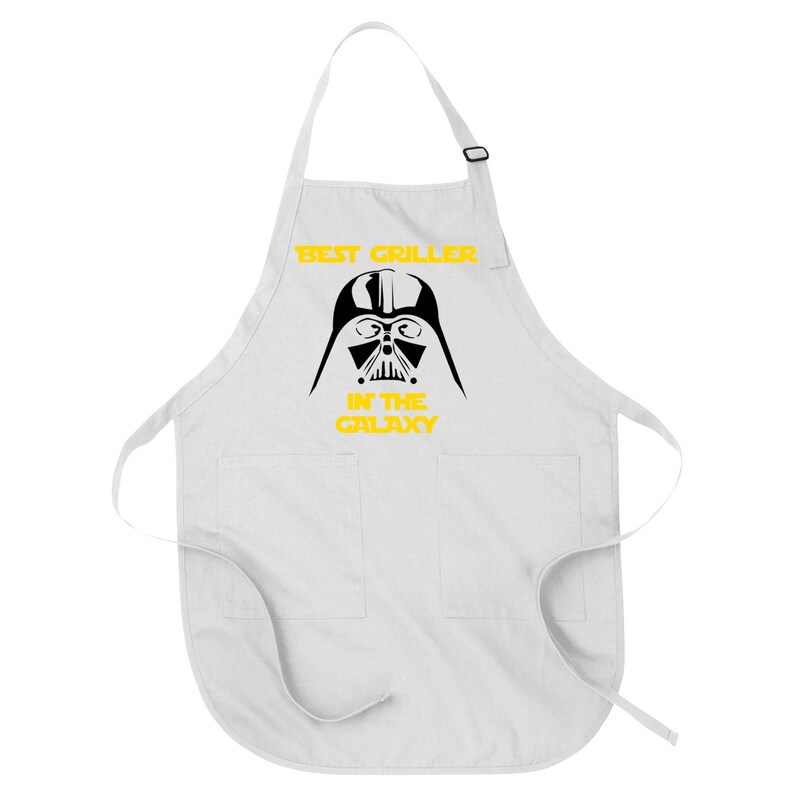 May include: White apron with two front pockets and an adjustable neck strap. The apron features a black graphic of Darth Vader's helmet and the text "BEST GRILLER IN THE GALAXY" in yellow.