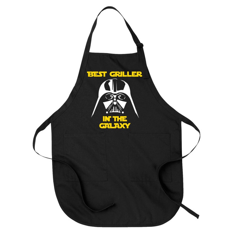 May include: Black apron with two front pockets and adjustable neck strap. The apron features a white graphic of Darth Vader's helmet and the text "BEST GRILLER IN THE GALAXY" in yellow, a Star Wars-themed design.