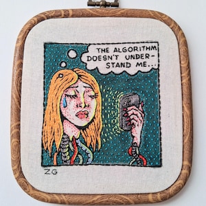 May include: A square embroidery hoop containing a fabric panel with a hand-stitched illustration of a person with blonde hair holding a phone. The text reads, THE ALGORITHM DOESN'T UNDERSTAND ME... The person has a blue tear and visible wires.