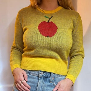Può includere: Un maglione giallo con un design a forma di mela rossa. Il maglione ha maniche lunghe e una trama a coste.