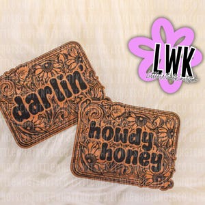 May include: Two rectangular wooden plaques with floral and scroll designs. One reads "darlin'" and the other "howdy honey" in black lettering. A pink flower-shaped logo with "LWK" is in the upper right corner.