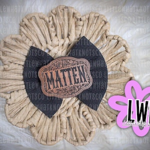 May include: A hair bow featuring a dark charcoal textured fabric base and a central copper-toned oval plaque with ornate filigree edges. The plaque is engraved with the name MATTEN in a bold, dark serif font, centered on a tan raffia flower.