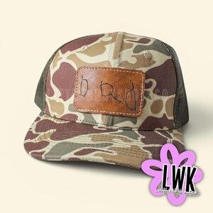 May include: A camouflage print trucker hat with a brown mesh back and a rectangular brown leather patch on the front. The patch features the handwritten word dad in black ink. The hat has a structured crown and a curved brim with a camo pattern.