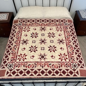 May include: A cream-coloured quilt with a star pattern in shades of burgundy and cream. The quilt features a border with leaf and berry designs. The bed frame is black, and the bed is made.