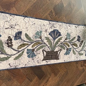 May include: A rectangular quilted table runner with a floral design. The runner has a cream background with sketched flowers. The central design shows a basket of blue and green flowers and leaves. It is trimmed with a dark blue border.