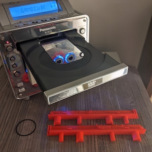 May include: A silver Panasonic SL-GC10 DVD player with a disc tray open, displaying a GameCube game. The screen reads "GameCube". Includes red plastic components and black rubber bands, likely for repair or replacement.