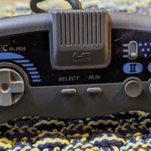 Könnte beinhalten: Grauer NEC PI-PD5-Gamecontroller mit Steuerkreuz, Tasten mit der Aufschrift „SELECT“ und „RUN“ sowie Tasten mit der Aufschrift „I“ und „II“. Der Controller hat blaue Akzente und ein schwarzes Kabel.