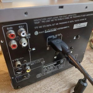 May include: A silver and black Panasonic SL-GC10 DVD/Game Player. The back panel features various input/output ports, including digital AV out, and text that reads "Manufactured under license from Dolby Laboratories". A black cable is plugged into the digital AV out port.