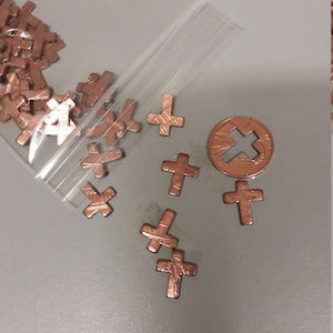 May include: A plastic bag filled with small copper-colored cross charms. The bag is open and the charms are spilling out. There is a single copper-colored penny with a cross design in the center of the image.