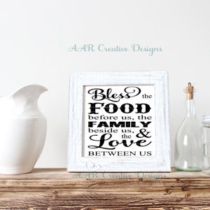 May include: White framed print with black text that reads "Bless the Food before us, the Family beside us, & the Love BETWEEN US".