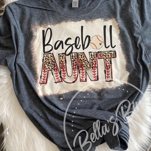 May include: A dark gray t-shirt with a white distressed background featuring the text "Baseball Aunt" in black with a leopard print design and a red baseball stitch design. A white baseball with a red heart is above the text.