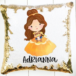 May include: Gold sequin pillow with a white front featuring a cartoon illustration of Belle from Beauty and the Beast holding a teddy bear. The pillow has the name "ADRIANNA" printed below the illustration.