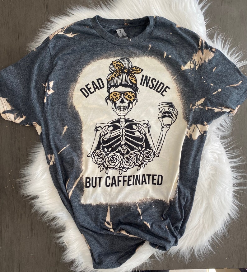 May include: A dark gray t-shirt with a bleached design featuring a skeleton holding a coffee cup. The text on the shirt reads "Dead Inside But Caffeinated".