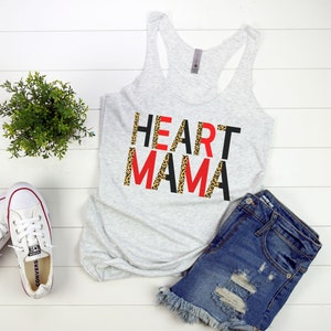 May include: A gray tank top with the words "HEART MAMA" printed in black, red, and leopard print. The tank top is laying on a white surface with a pair of white sneakers and denim shorts.