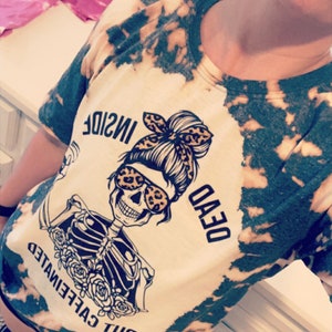 May include: A white t-shirt with a tie-dye pattern in black and green. The shirt features a graphic of a skeleton wearing sunglasses and a bun with a bandana. The text on the shirt reads "BUT CAFFEINEATED DEAD INSIDE".