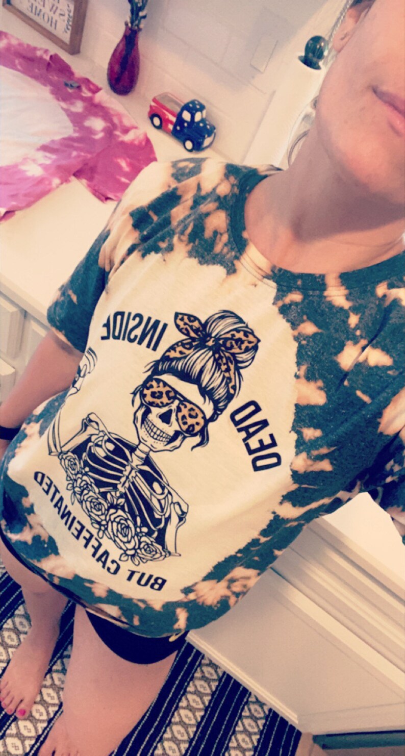 May include: A white t-shirt with a tie-dye pattern in black and green. The shirt features a graphic of a skeleton wearing sunglasses and a bun with a bandana. The text on the shirt reads "BUT CAFFEINEATED DEAD INSIDE".