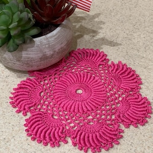 May include: A pink crocheted doily with a circular design in the center. The doily is on a light brown countertop.