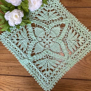 May include: A square, mint green crocheted doily with a floral and leaf pattern. The doily is placed on a wooden surface and is decorated with white roses and green leaves.
