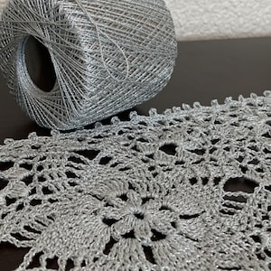 May include: A close-up of a silver crochet lace and a ball of matching yarn. The lace features a floral design with intricate openwork. The yarn has a metallic sheen, suggesting a decorative or craft project.