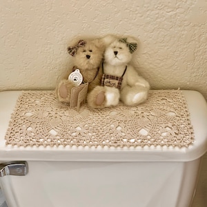 May include: Two beige teddy bears with brown eyes and brown noses are sitting on a crocheted beige doily. The bear on the left has a brown bow and the bear on the right has a green bow. The bear on the right is wearing a brown and white vest with the word "Friends" on it. The doily is on top of a white toilet tank.
