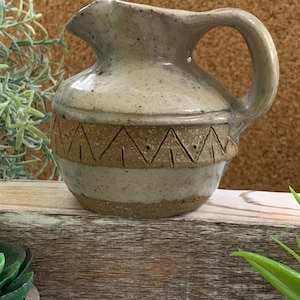 May include: A small, gray ceramic pitcher with a brown, geometric design around the middle. The pitcher has a handle and a slightly flared lip.
