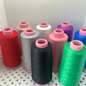 May include: Assortment of spools of thread in various colors, including red, black, green, blue, pink, white, gray, and beige. The spools are arranged in a semi-circle on a white surface.