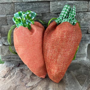 May include: Two carrot-shaped burlap bags with green and white checkered fabric tops and green ribbon ties. The bags are orange and sit on a stone surface against a gray stone wall.