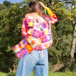 May include: A woman wearing a colorful crocheted sweater with a patchwork design. The sweater is made of different colored squares with flowers in each square. The woman is wearing blue jeans and has long brown hair.