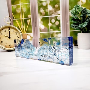 May include: A clear acrylic desk nameplate with a floral design in shades of blue and white. The nameplate features the name "Ashlyn Kennedy" and the title "MD INTERNAL MEDICINE" in black script.