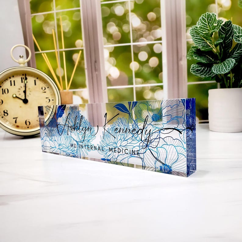 May include: A clear acrylic desk nameplate with a floral design in shades of blue and white. The nameplate features the name "Ashlyn Kennedy" and the title "MD INTERNAL MEDICINE" in black script.