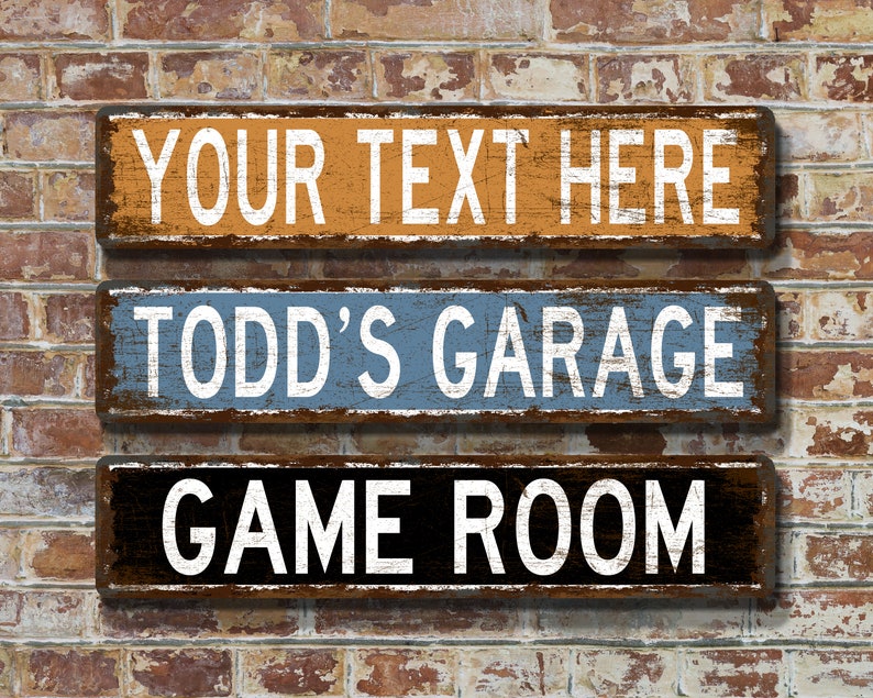 May include: Three rectangular wooden signs with white text against a distressed background. The top sign is orange and reads "YOUR TEXT HERE." The middle sign is blue and says "TODD'S GARAGE." The bottom sign is black and says "GAME ROOM."