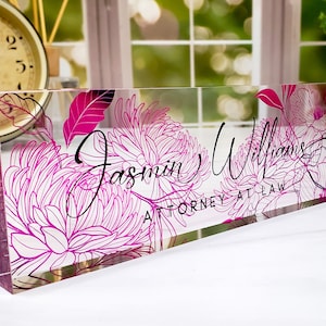 May include: A clear acrylic desk nameplate featuring a pink floral design. The name "Jasmin Williams" and the title "ATTORNEY AT LAW" are written in black script. Ideal for a professional setting.
