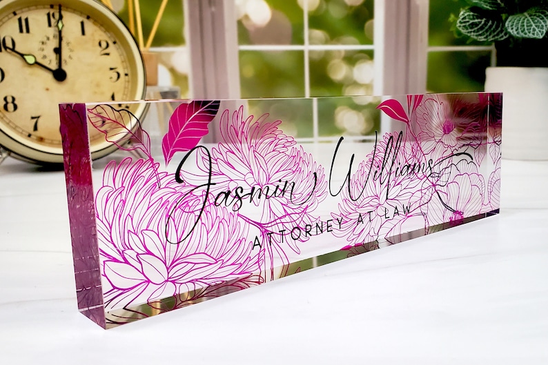 May include: A clear acrylic desk nameplate featuring a pink floral design. The name "Jasmin Williams" and the title "ATTORNEY AT LAW" are written in black script. Ideal for a professional setting.