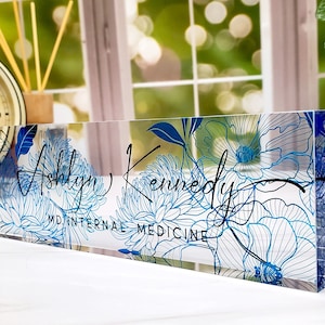 May include: A clear acrylic desk nameplate featuring a floral design in shades of blue. The name "Ashlyn Kennedy" is written in elegant script, with "MD INTERNAL MEDICINE" below. The nameplate is rectangular and stands upright.
